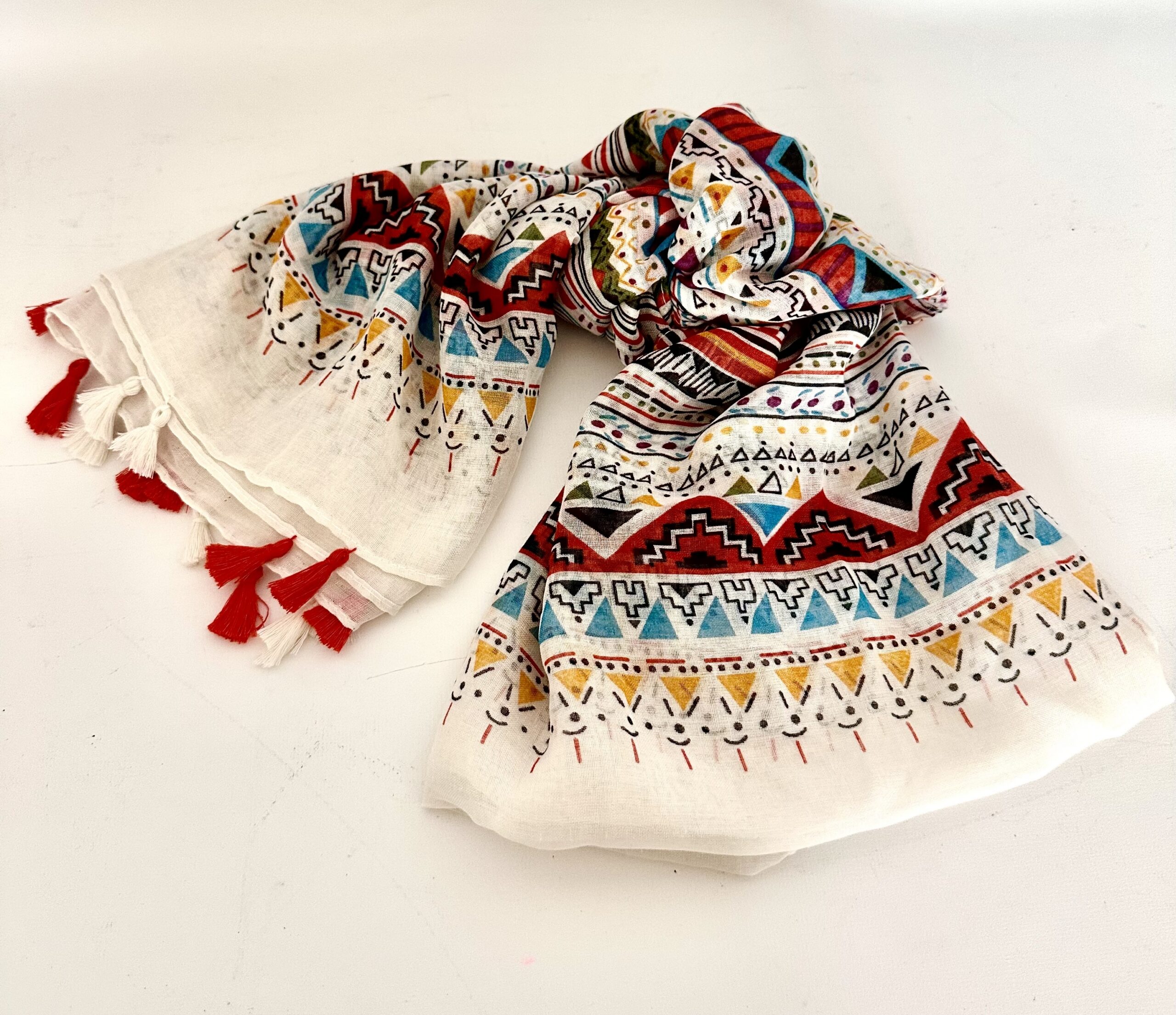 Lightweight cream scarf printed with bands of geometric patterns in terracotta red, turquoise, mustard yellow and black, finished with alternating red and white tassels along the ends.