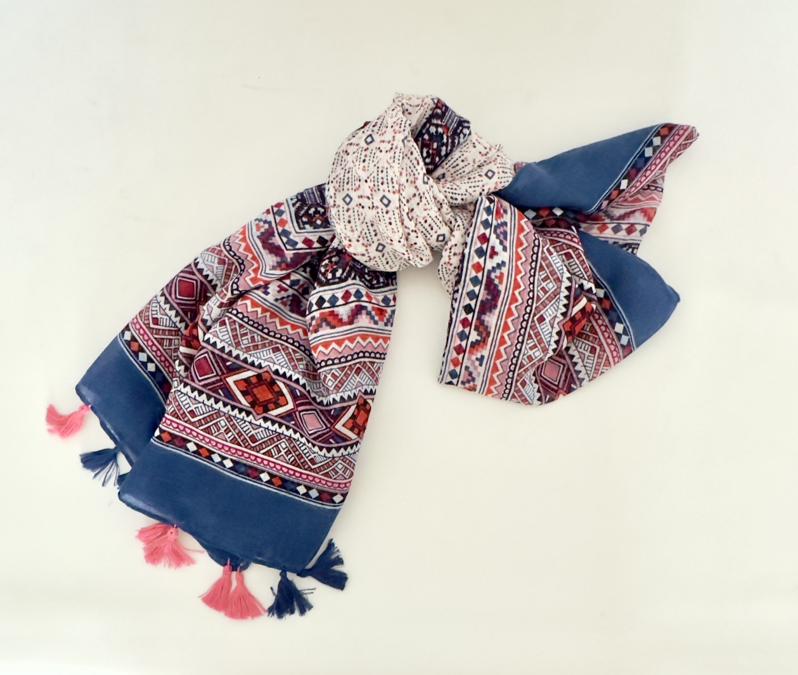Lightweight scarf with wide solid blue ends, bands of geometric diamonds and zigzags in coral, pink, burgundy and navy, and a cream centre dotted with small diamonds, finished with alternating pink and navy tassels.