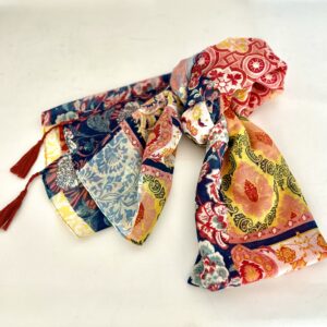 Lightweight scarf printed in a patchwork of floral and tile patterns in navy, sky blue, sunny yellow, coral pink and red, with long rust-coloured corded tassels at the corners.