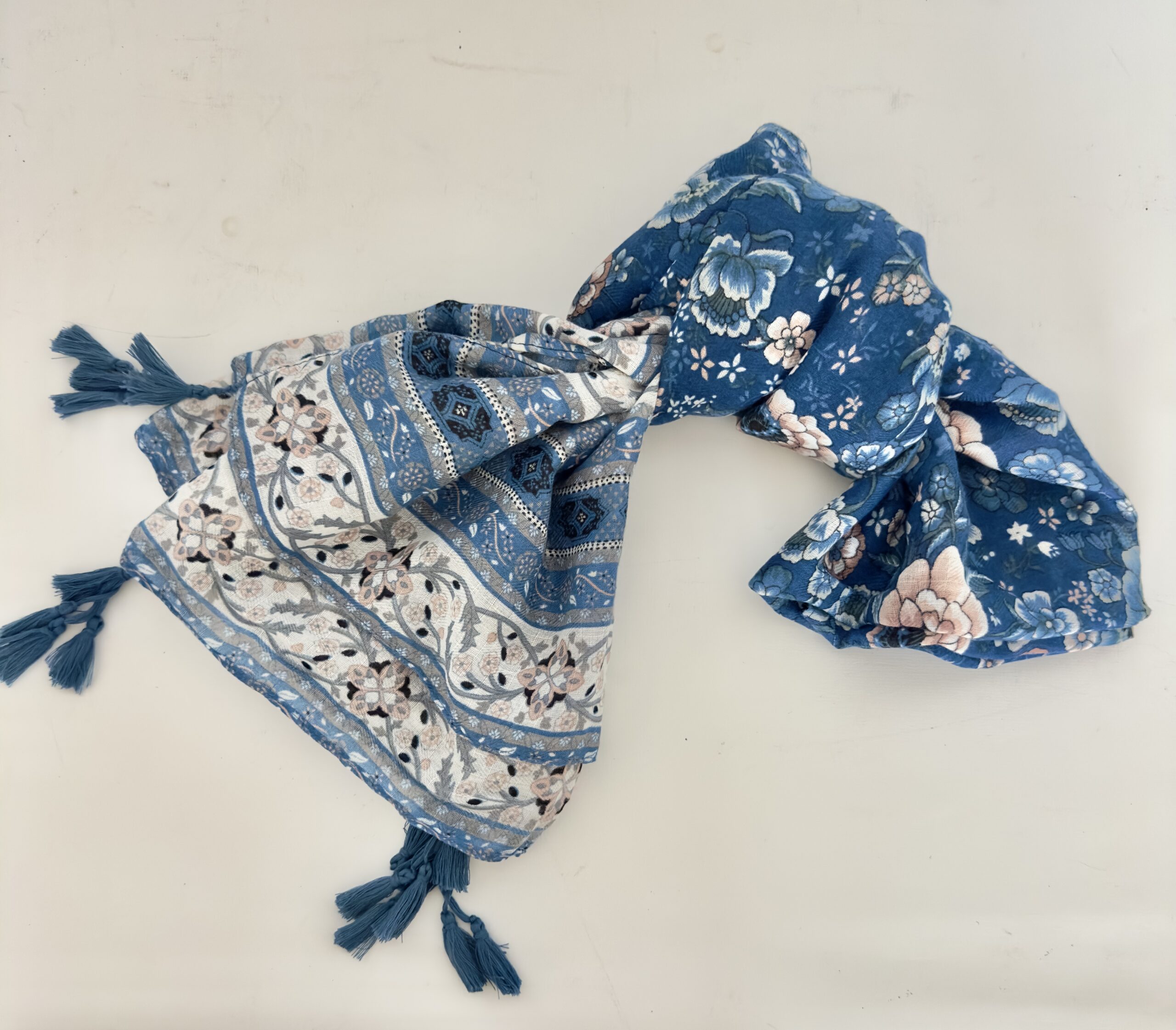 Lightweight scarf in denim blue with large blush pink, white and pale blue flowers, and ends bordered with bands of cream florals, navy medallions and dotted stripes, finished with clusters of blue tassels at the corners.