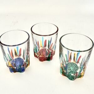 Three clear shot glasses with star-shaped bases in blue, ruby red and green, each decorated with pointed rays of red, orange, yellow, turquoise, blue and green rising from the base.