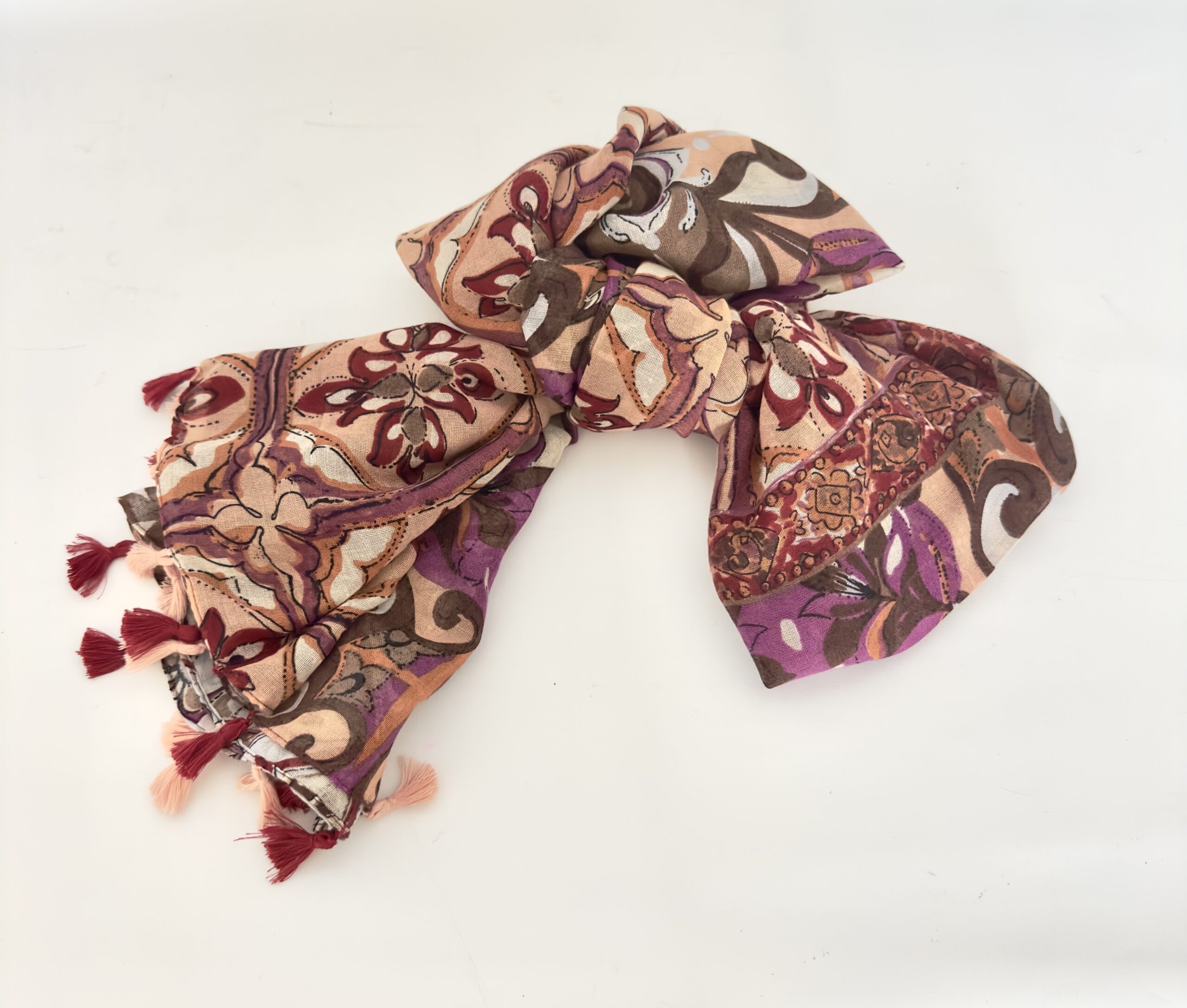 Lightweight scarf printed with large swirling florals and medallions in peach, plum purple, burgundy, chocolate brown and cream, finished with alternating burgundy and peach tassels.